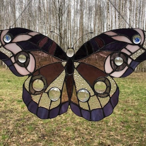 May include: A stained glass butterfly with purple, brown, and white accents. The butterfly has clear glass circles throughout the wings.