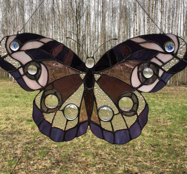 May include: A stained glass butterfly with purple, brown, and white accents. The butterfly has clear glass circles throughout the wings.