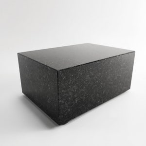 Puede incluir: Un bloque rectangular de granito negro con una superficie superior pulida. El bloque tiene un aspecto moteado y est&aacute; sobre un fondo blanco. El dise&ntilde;o es minimalista, adecuado para exhibici&oacute;n o como base, con unos 30 cm de largo.