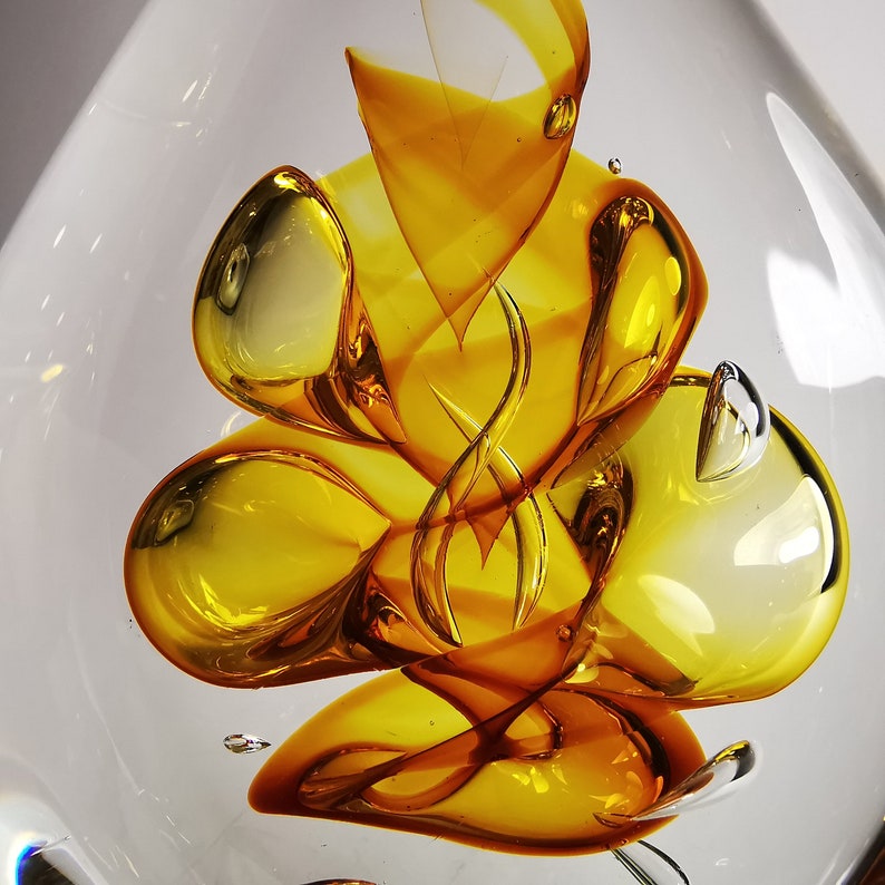 May include: A clear glass sculpture featuring internal swirling patterns of amber and golden yellow hues. The design includes organic, rounded shapes and elongated, twisted ribbons of color suspended within the transparent glass body.