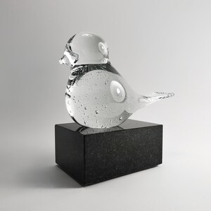 Puede incluir: Una escultura de p&aacute;jaro de cristal transparente con burbujas de aire, colocada sobre un bloque de granito negro. El p&aacute;jaro tiene una forma redondeada y un pico peque&ntilde;o. Es un objeto decorativo.