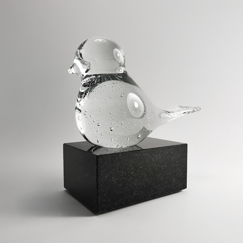 Puede incluir: Una escultura de p&aacute;jaro de cristal transparente con burbujas de aire, colocada sobre un bloque de granito negro. El p&aacute;jaro tiene una forma redondeada y un pico peque&ntilde;o. Es un objeto decorativo.