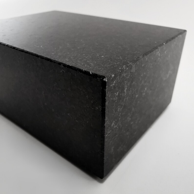 Puede incluir: Un bloque rectangular negro s&oacute;lido con una superficie texturizada. El bloque tiene un lado ligeramente inclinado, creando una forma geom&eacute;trica. El objeto est&aacute; hecho de un material oscuro, posiblemente piedra o una sustancia similar.