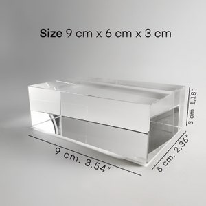 Könnte beinhalten: Ein transparenter Acrylblock mit den Maßen 9 cm x 6 cm x 3 cm. Der Block ist rechteckig und hat eine glatte, polierte Oberfläche.