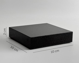 Puede incluir: Un bloque cuadrado negro de 10 cm x 10 cm y 2 cm de grosor. El bloque tiene un acabado mate y una superficie ligeramente texturizada. Podría ser un soporte de exhibición o una pequeña plataforma.