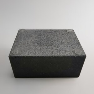 Puede incluir: Un bloque rectangular de piedra gris oscuro con una superficie texturizada. El bloque tiene cuatro peque&ntilde;os pies circulares transparentes adheridos en la parte inferior. La piedra tiene un aspecto moteado y est&aacute; sobre un fondo blanco.