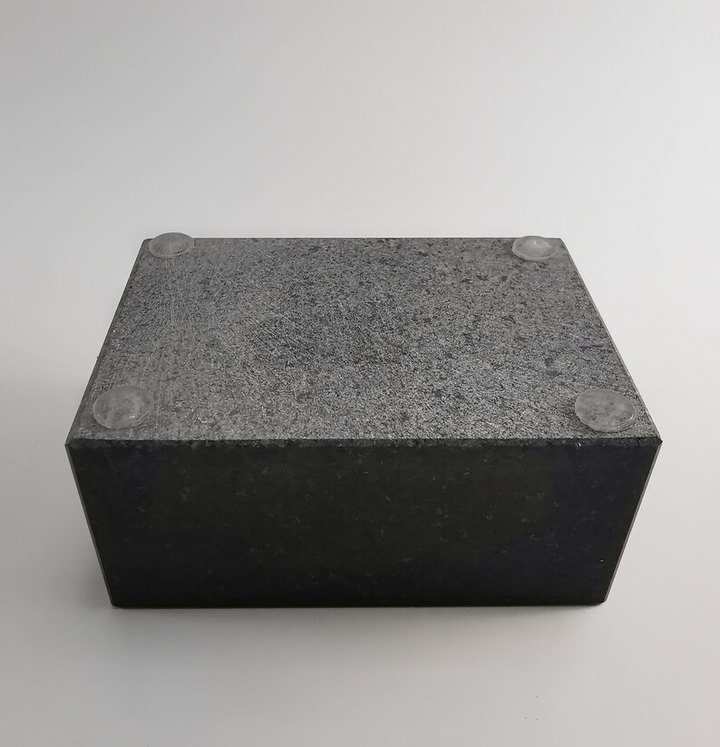 Puede incluir: Un bloque rectangular de piedra gris oscuro con una superficie texturizada. El bloque tiene cuatro peque&ntilde;os pies circulares transparentes adheridos en la parte inferior. La piedra tiene un aspecto moteado y est&aacute; sobre un fondo blanco.