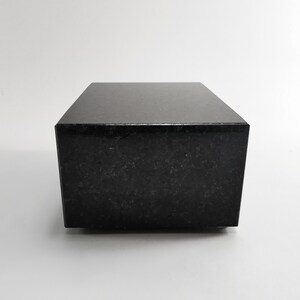 Puede incluir: Un bloque rectangular negro s&oacute;lido con una superficie pulida y reflectante. El bloque tiene un patr&oacute;n moteado sutil y est&aacute; sobre un fondo blanco liso. El objeto parece estar hecho de piedra o un material similar.