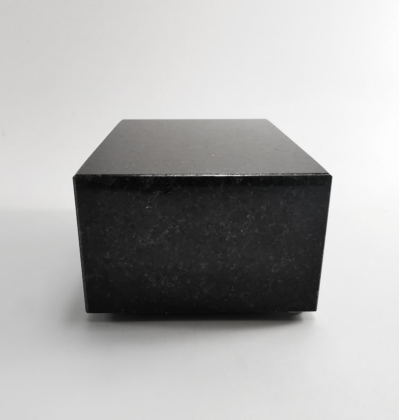 Puede incluir: Un bloque rectangular negro s&oacute;lido con una superficie pulida y reflectante. El bloque tiene un patr&oacute;n moteado sutil y est&aacute; sobre un fondo blanco liso. El objeto parece estar hecho de piedra o un material similar.