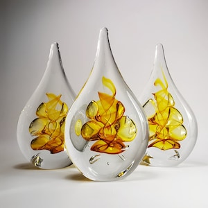 May include: Three teardrop-shaped glass sculptures with clear outer layers and internal swirling patterns of vibrant yellow and orange glass. Each piece features a smooth, rounded base that tapers to a point at the top, showcasing intricate glasswork.