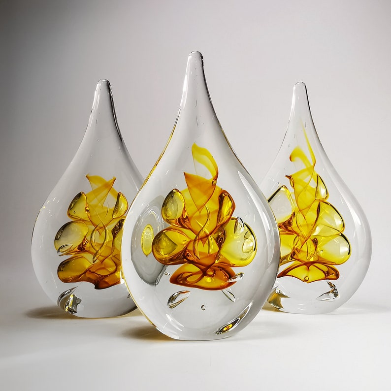 May include: Three teardrop-shaped glass sculptures with clear outer layers and internal swirling patterns of vibrant yellow and orange glass. Each piece features a smooth, rounded base that tapers to a point at the top, showcasing intricate glasswork.