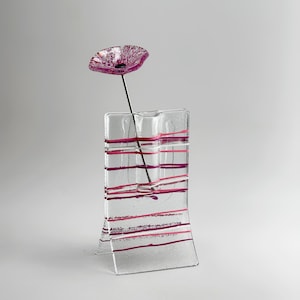 May include: A clear glass vase with a rectangular, curved base and horizontal pink and purple stripes. A single decorative glass flower with a dark purple center and light purple petals sits inside the vase on a thin, dark metal stem.