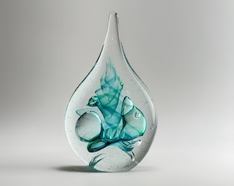 May include: A teardrop-shaped clear glass sculpture featuring an internal swirling pattern of teal and blue hues. The solid glass piece contains small air bubbles suspended throughout the clear material, creating a textured and layered visual effect.