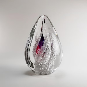 Może przedstawiać: Szklany, przezroczysty przycisk do papieru w kształcie łzy z wirem czerwonego i fioletowego szkła w środku. Szkło jest wypełnione małymi pęcherzykami powietrza.