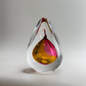 Könnte beinhalten: Ein tropfenförmiger Briefbeschwerer aus Glas mit einer klaren Außenschicht und einem inneren Kern in einer leuchtenden Mischung aus Magenta und Bernstein. Das Innere enthält kleine Luftblasen und ein stielartiges Detail oben in der Mitte.