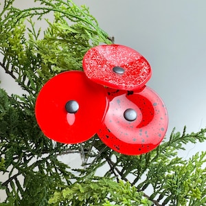 Könnte beinhalten: Drei kreisförmige Glasornamente in leuchtendem Rot, jeweils mit einem kleinen dunkelgrauen Mittelknopf. Ein Ornament hat eine einfarbige rote Oberfläche, ein weiteres eine gesprenkelte weiße Textur und das dritte ein schwarzes Spritzmuster.