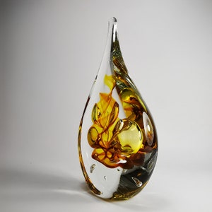 May include: A teardrop-shaped glass sculpture featuring clear glass encasing swirling patterns of amber, yellow, and brown hues. The solid object has a smooth, polished surface with visible internal air bubbles and a pointed top.