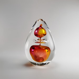 Op de afbeelding: Een transparante glazen sculptuur in de vorm van een traan met een rode en oranje glazen bol erin. De bol hangt in het midden van de sculptuur en lijkt te zweven.