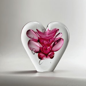 Può includere: Un fermacarte a forma di cuore in vetro trasparente con un vivace disegno floreale rosa all'interno. Il design rosa ha un aspetto astratto a strati. Il fermacarte è esposto su una superficie bianca.