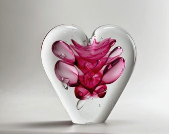 Puede incluir: Un pisapapeles en forma de corazón de cristal transparente con un vibrante diseño floral rosa en el interior. El diseño rosa tiene una apariencia abstracta en capas. El pisapapeles se exhibe sobre una superficie blanca.