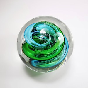 May include: A clear glass paperweight with a swirling design of teal and green. The colors are swirled together in a spiral pattern, creating a dynamic and eye-catching effect. Bubbles are suspended within the glass.