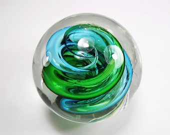 May include: A clear glass paperweight with a swirling design of teal and green. The colours are swirled together in a spiral pattern, creating a dynamic effect. Bubbles are suspended within the glass.