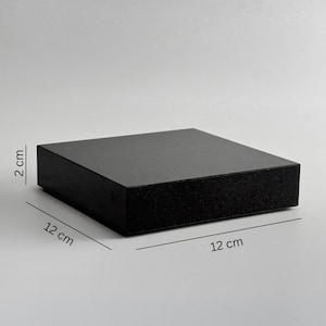 Puede incluir: Un bloque cuadrado negro, de 12 cm x 12 cm y 2 cm de grosor. El bloque tiene un acabado mate y se muestra sobre un fondo blanco liso. Las dimensiones están claramente indicadas en centímetros.