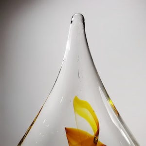 May include: A clear, conical glass sculpture featuring a pointed top and a smooth, transparent surface. Inside the glass, there is a swirling, abstract pattern of vibrant yellow and orange hues, with small air bubbles visible throughout the structure.