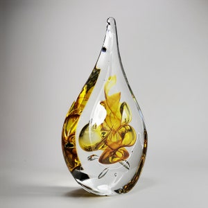 May include: A teardrop-shaped glass sculpture featuring a clear exterior shell and an internal core of swirling amber and yellow glass. The piece has a smooth, polished surface with visible air bubbles trapped within the clear glass structure.