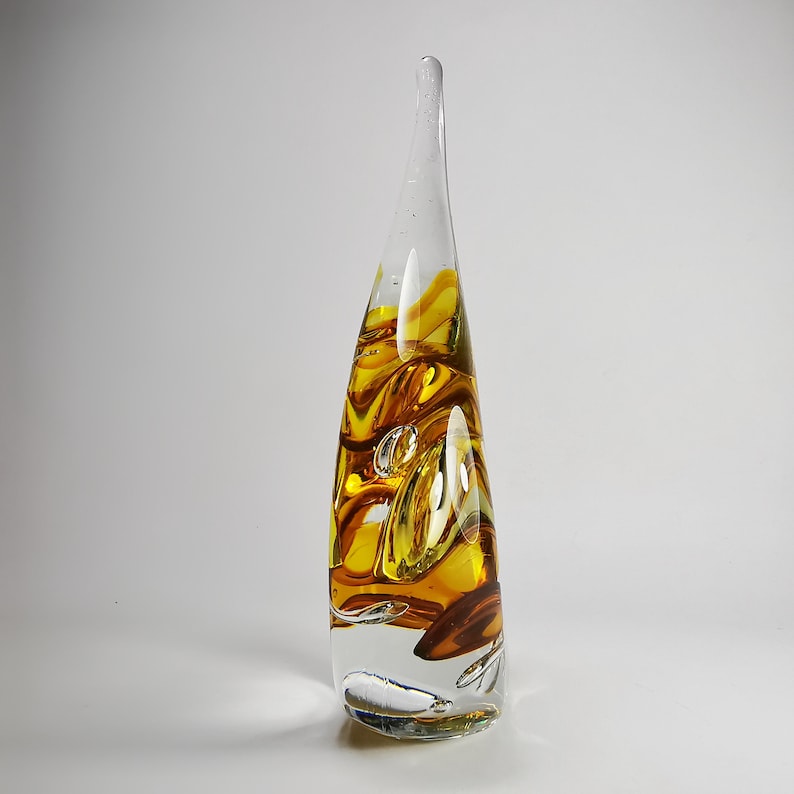 May include: A tall, teardrop-shaped glass sculpture featuring a clear base that transitions into swirling amber and golden-yellow hues. The interior contains suspended air bubbles, and the elongated form tapers to a smooth, rounded point at the top.