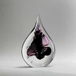 Puede incluir: Una escultura de vidrio en forma de lágrima con una capa exterior transparente y un núcleo interno de remolinos de vidrio en tonos violeta oscuro, negro y violeta translúcido. La pieza sólida tiene una superficie lisa y pulida con una punta.