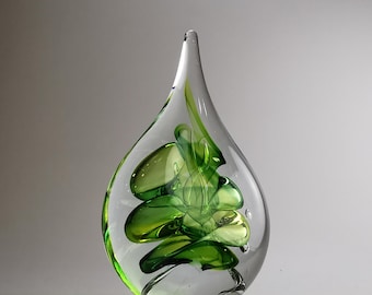 May include: A teardrop-shaped glass sculpture featuring a clear outer layer and an internal swirling design of vibrant green glass. The piece has a smooth, polished surface and a pointed top, with small air bubbles visible within the glass structure.