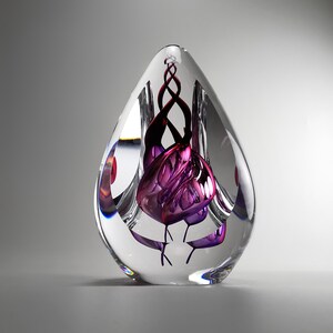 Può includere: Un fermacarte in vetro trasparente a forma di goccia contenente una scultura interna di nastri di vetro viola e rosso scuro intrecciati. L'oggetto in vetro massiccio ha una superficie liscia e lucida che rifrange la luce, mettendo in risalto i complessi motivi intrecciati sospesi nel nucleo trasparente.