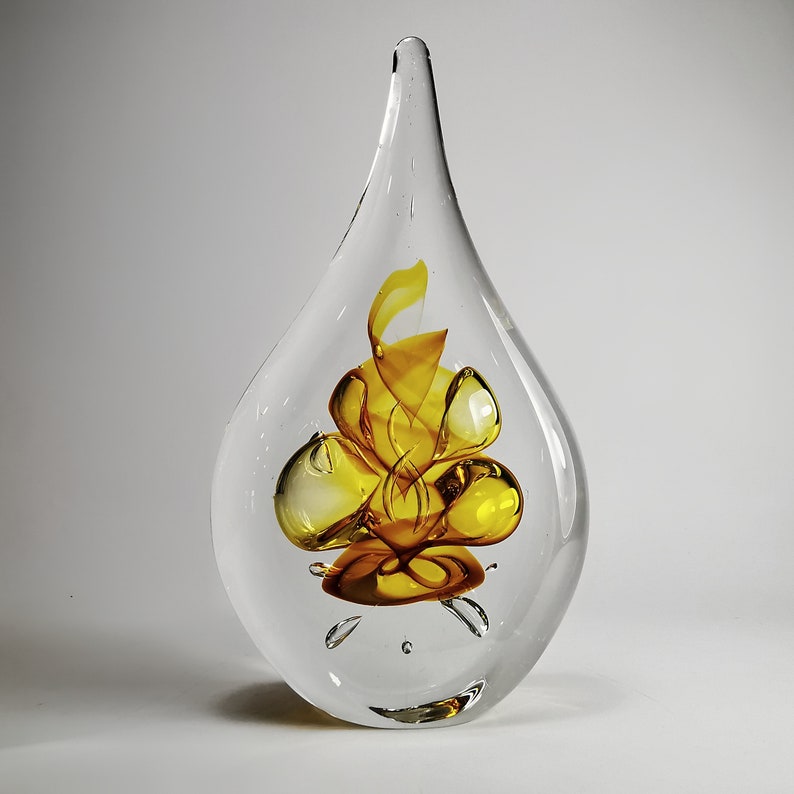 May include: A clear, teardrop-shaped glass sculpture featuring an internal, swirling amber and yellow glass design. The object has a smooth, polished exterior surface and a pointed top, with the vibrant colored glass suspended within the center.