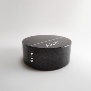 May include: A dark, cylindrical object with a speckled texture. The top surface is flat and round, measuring 9.5 cm in diameter. The height of the cylinder is 4 cm. The object is likely made of stone.