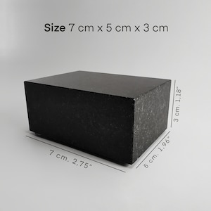 Könnte beinhalten: Ein massiver schwarzer rechteckiger Block, 7 cm x 5 cm x 3 cm, mit strukturierter Oberfläche. Die Abmessungen sind auf dem Bild angegeben. Der Block besteht aus dunklem Material und liegt auf einer hellgrauen Oberfläche.