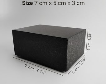 Puede incluir: Un bloque rectangular negro macizo, de 7 cm x 5 cm x 3 cm, con una superficie texturizada. Las dimensiones están etiquetadas en la imagen. El bloque está hecho de un material oscuro y se asienta sobre una superficie gris claro.
