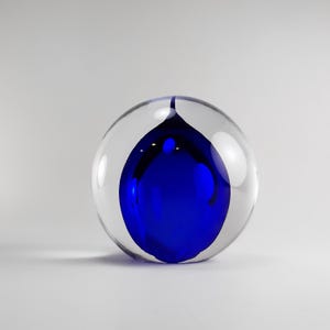 Könnte beinhalten: Eine klare Glaskugel mit einer blauen Glaskugel im Inneren. Die blaue Kugel hat eine Tropfenform und ist teilweise mit Luftblasen gefüllt.