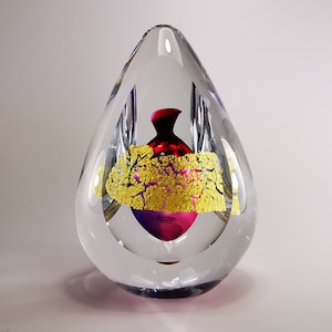 May include: Clear glass paperweight in a teardrop form. A red and purple vase is encased within, adorned with gold leaf. This decorative art piece would suit any home.