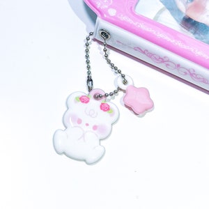 May include: A white bear charm with pink roses on its head, attached to a silver chain with a pink star charm.
