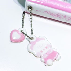 May include: A pink and white charm with a bear wearing a pink dress and a heart charm attached to a silver chain.