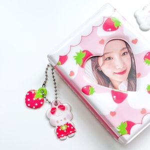 May include: A pink photo album with a heart-shaped window and a strawberry pattern. The album has a silver chain with a strawberry and a bear charm.