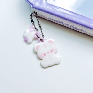 May include: A white and pink charm with a cat design attached to a silver chain. The charm is hanging from a purple and white patterned object.