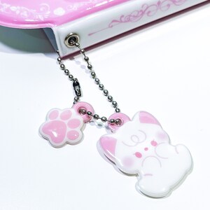 May include: A pink and white notebook with a silver chain attached to a pink paw print charm and a white cat charm.