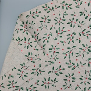 Può includere: Un tessuto beige con un motivo ripetuto di foglie di agrifoglio verdi e bacche rosse.