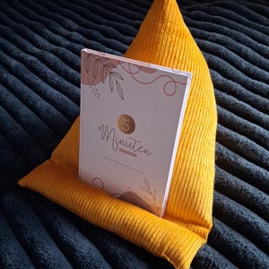May include: A yellow corduroy book holder with a book titled "5 Minuten" resting inside. The book has a white cover with floral and line art designs. The book holder is on a dark blue textured surface.