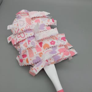 Può includere: Un piumino rosa e bianco con manico bianco e base rosa. La testa del piumino è realizzata in tessuto a strati e arricciato con un motivo di cuori, fiori e pois rosa, viola e arancioni.