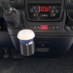 Könnte beinhalten: Schwarzer Auto-Innenraum mit einer Kaffeetasse in einem Getränkehalter. Der Becher ist blau und weiß mit einem weißen Deckel. Das Armaturenbrett hat ein digitales Display und verschiedene Knöpfe. Schlüssel hängen.