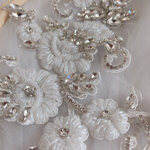 May include: Close-up of a white beaded and rhinestone floral applique. The applique features delicate white beads and sparkling rhinestones arranged in a floral pattern.