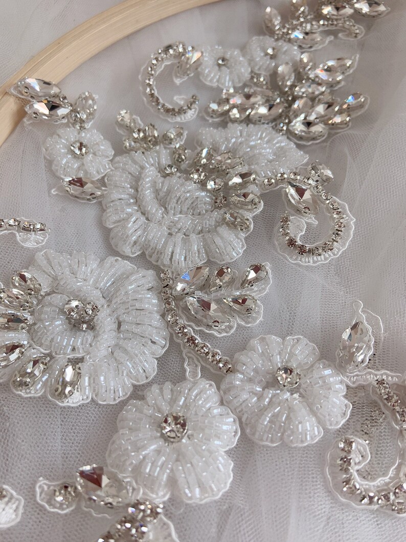 May include: Close-up of a white beaded and rhinestone floral applique. The applique features delicate white beads and sparkling rhinestones arranged in a floral pattern.